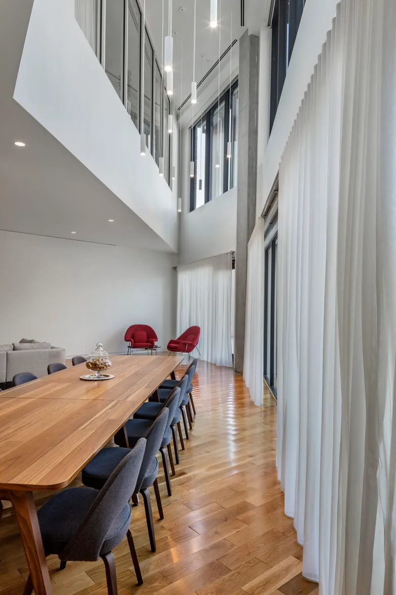 Double-height dining room with long timber table beside floor-to-ceiling sheer curtains and chevron wood flooring