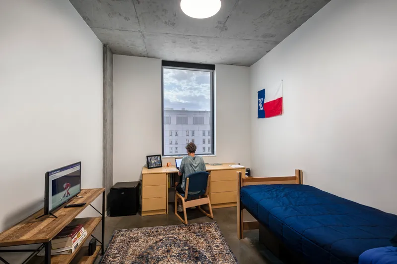 Dormitory room with exposed concrete ceiling, narrow window framing cloudy sky and neighboring buildings