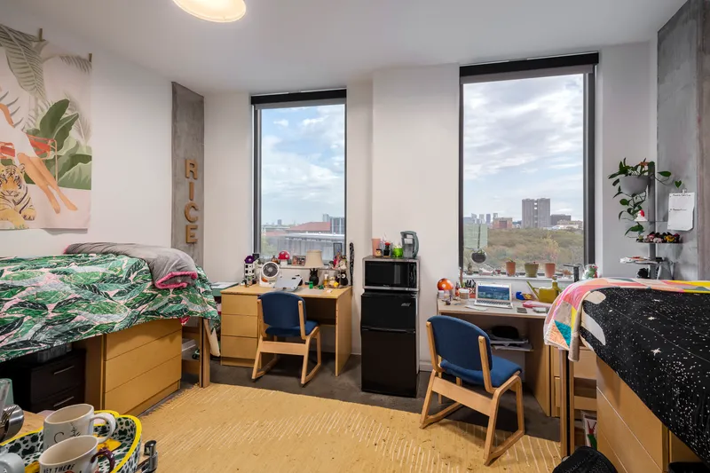 Shared dormitory room with corner windows overlooking the city skyline under partly cloudy skies