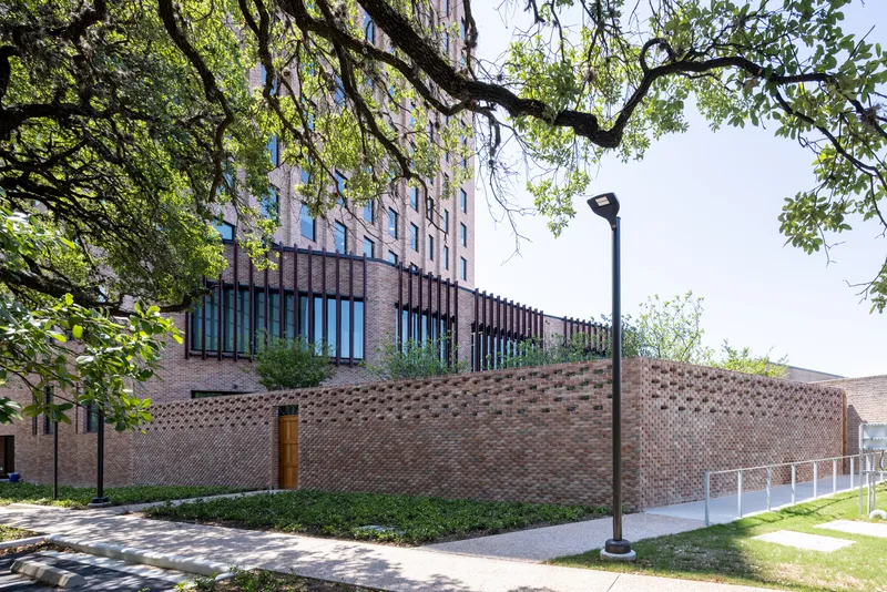 Perforated brick screen wall below a vertical tower with overhanging tree branches framing the view