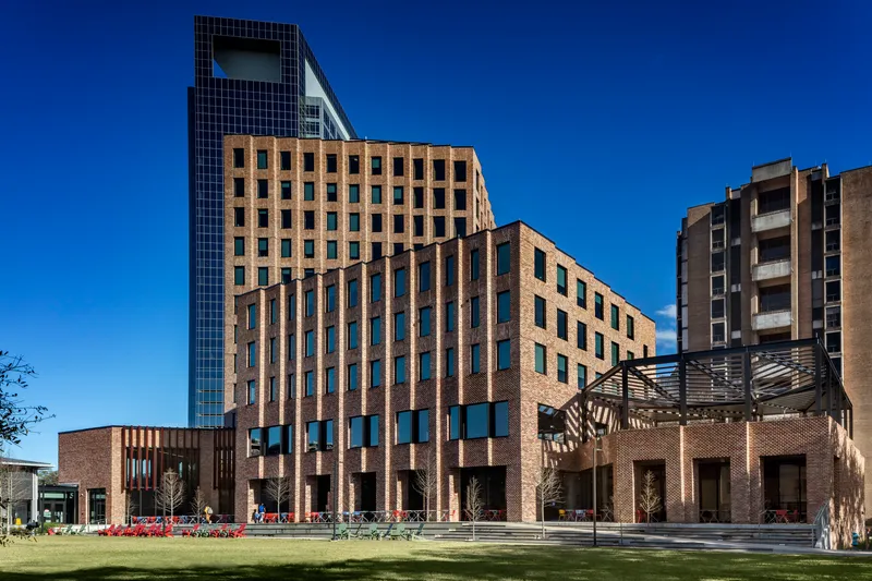 Brick tower with punched windows and adjacent black glass volume rising above a two-story base