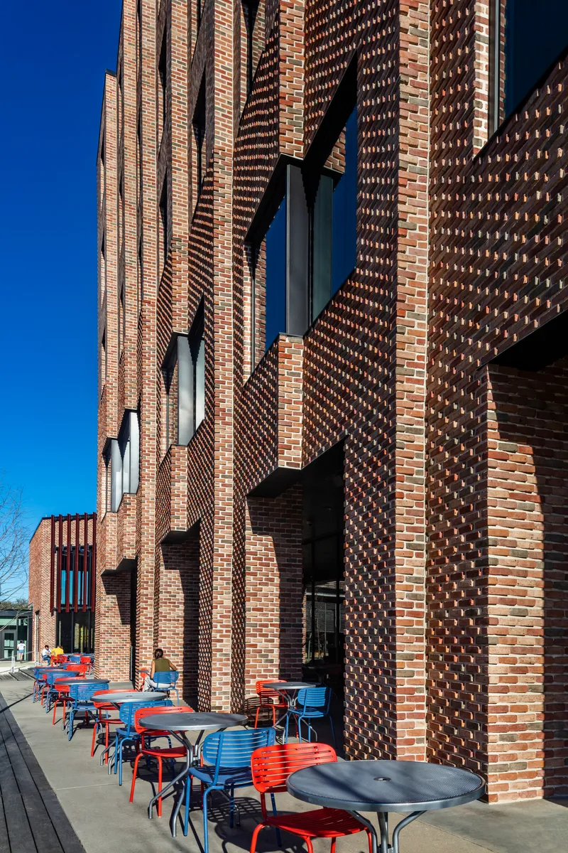 Textured brick facade with protruding and recessed patterns beside outdoor seating area with red and blue chairs