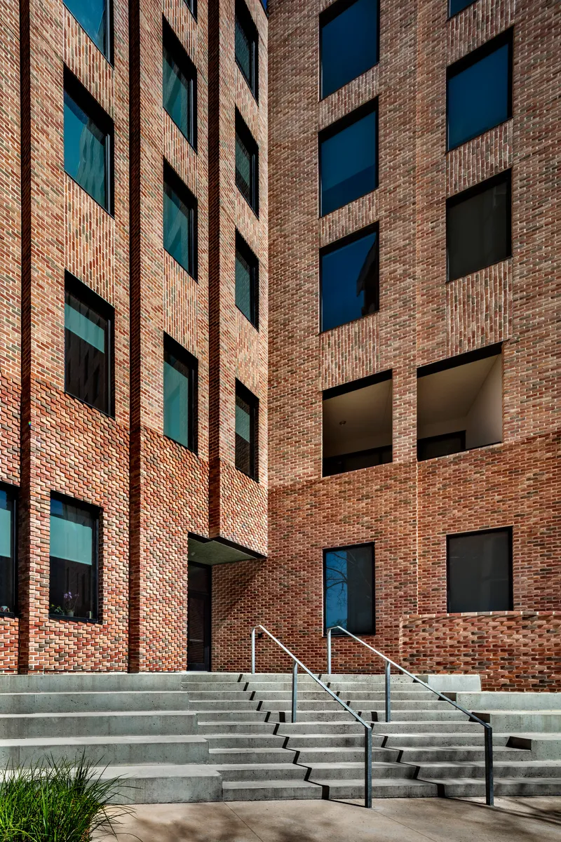 Concrete steps with metal handrails leading up to brick facade with punched window openings in afternoon sun