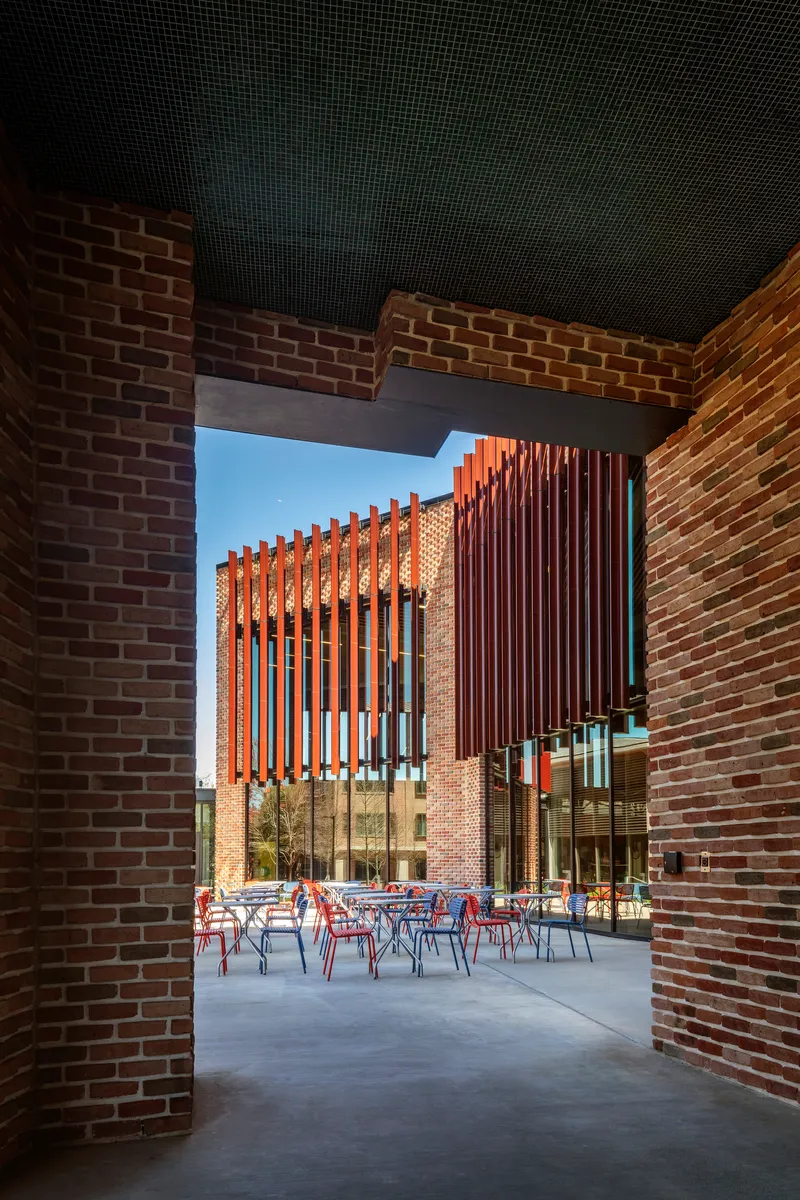 View through a brick-walled passageway into a courtyard with red chairs and vertical timber louvres beyond