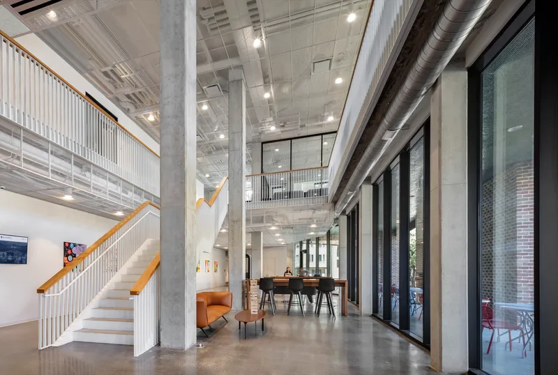 Double-height interior lobby with exposed concrete columns, a white steel staircase, and polished concrete floors