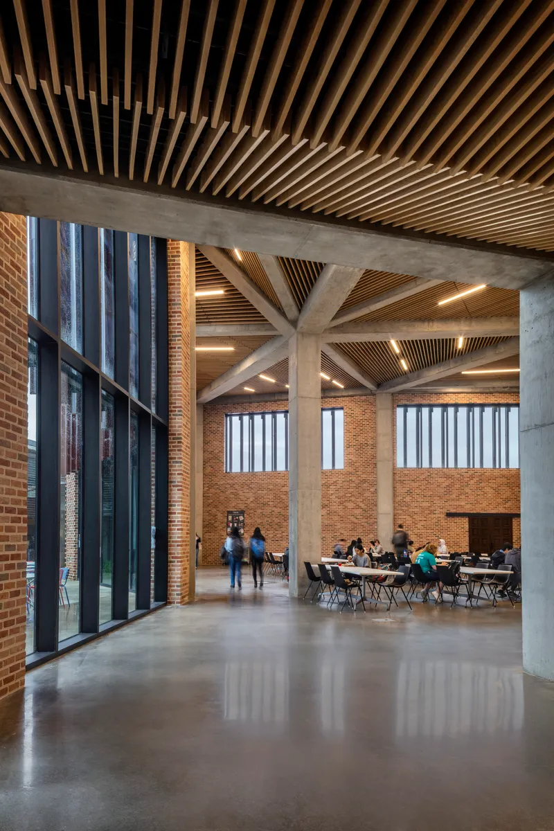 Concrete columns supporting a timber baffle ceiling above a polished floor with people seated in the background