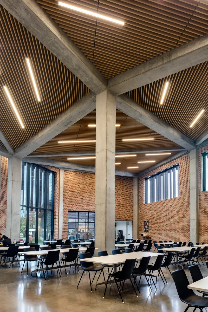 Cafeteria space with diagonal concrete beams spanning the timber slat ceiling and linear light fixtures between them