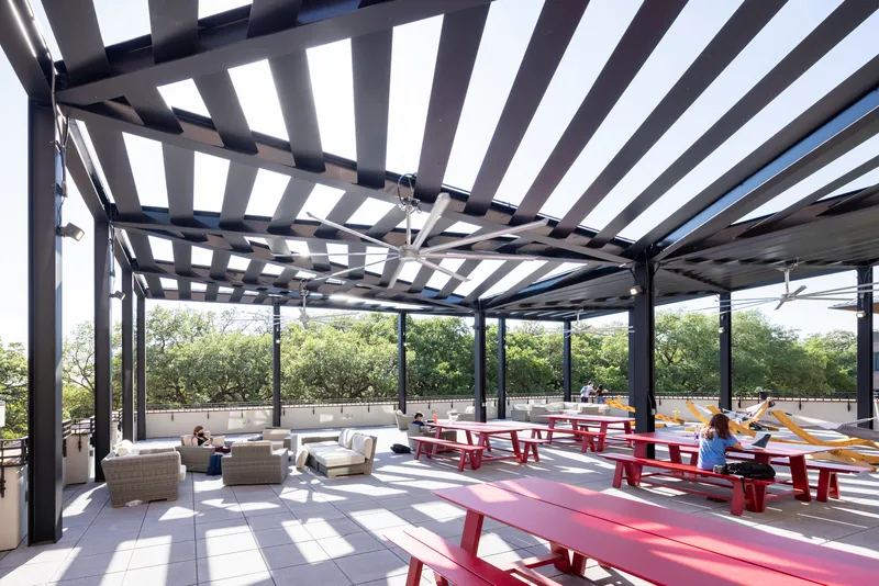 Rooftop terrace with open steel pergola slats casting shadows across red picnic tables under midday sun