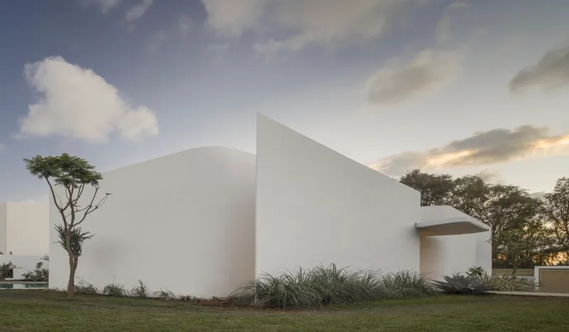 White stucco facade with curved opening flanked by grasses and a tree at dusk