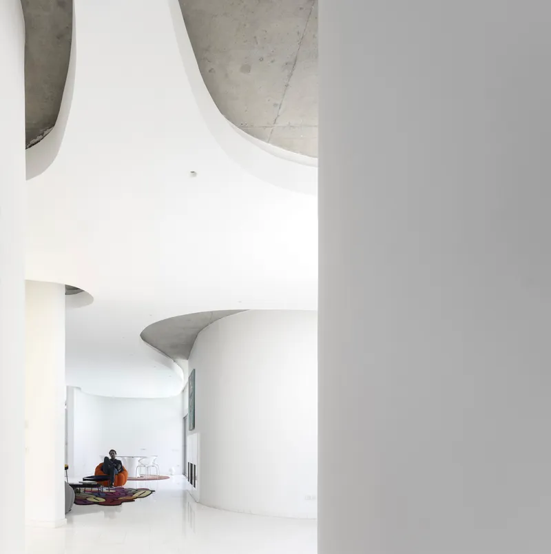 Curved white wall meeting a sculptural concrete ceiling element with a seated figure in the background