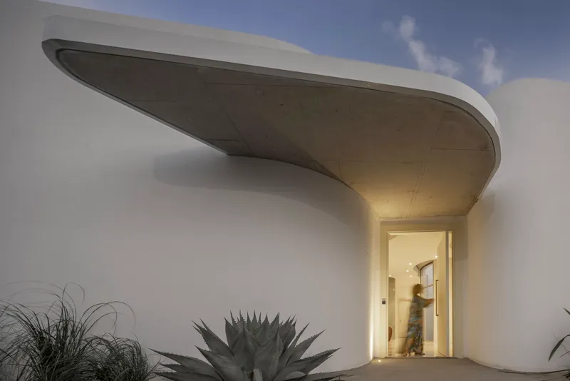 Illuminated entrance beneath a curved concrete canopy with agave and grasses at dusk