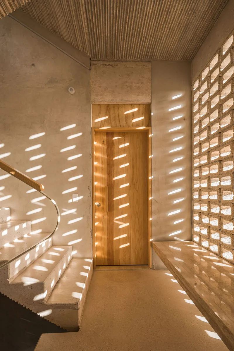 Entry foyer with perforated brick screen filtering dappled sunlight across concrete stairs and timber slatted ceiling
