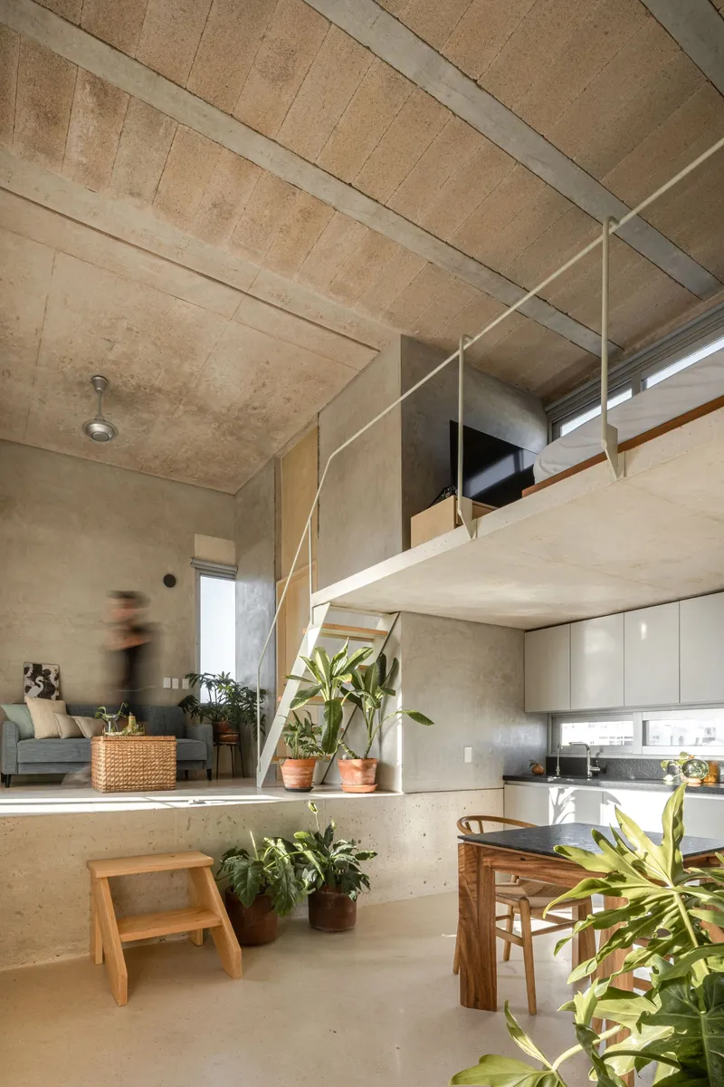 Double-height living space with exposed concrete ceiling, mezzanine level, and person in motion near potted plants