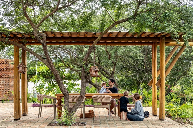 Timber pergola with exposed rafters sheltering a dining table and benches beneath mature trees