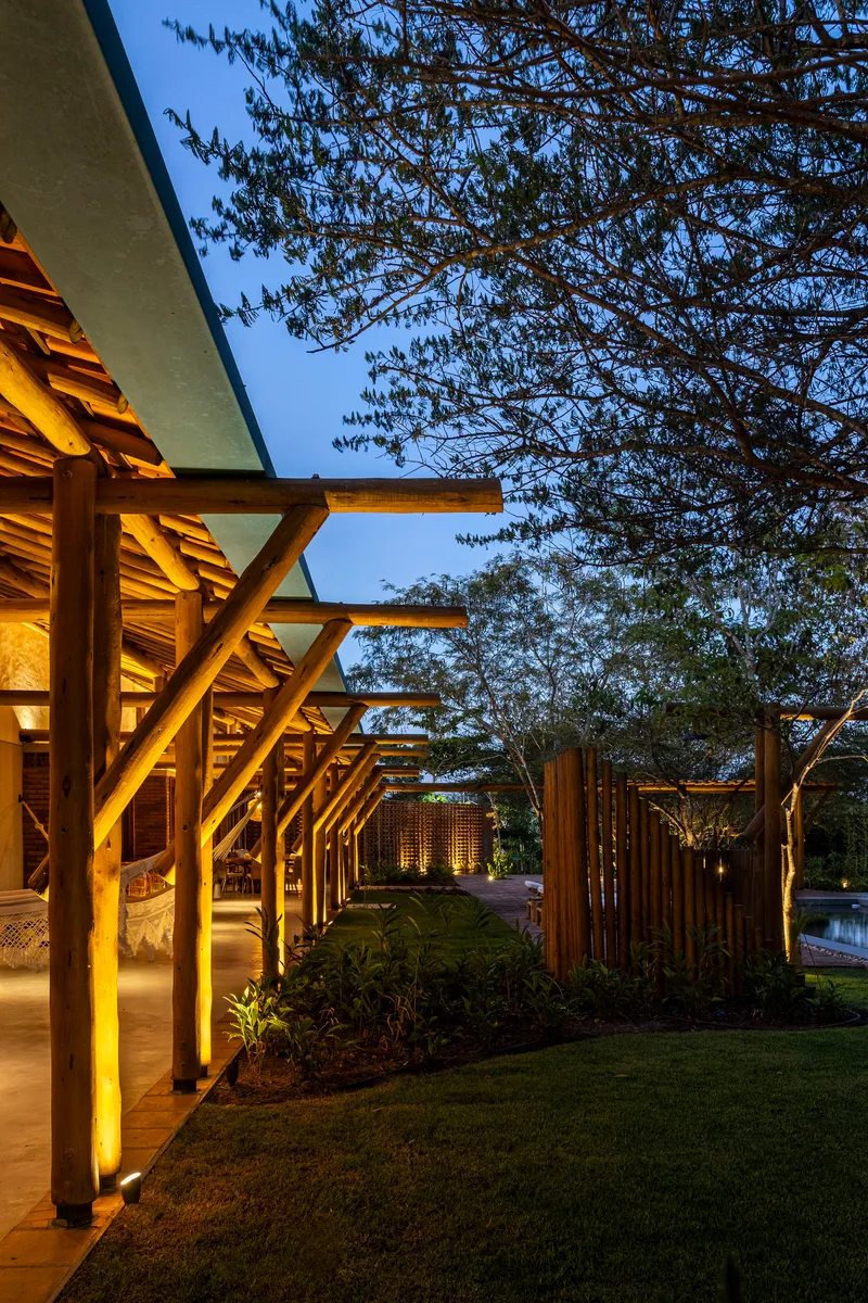 Illuminated timber colonnade with exposed beams supporting a flat roof beside lawn and mature trees at dusk