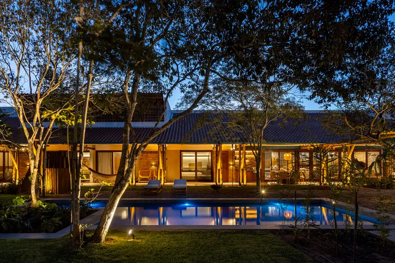 Tiled roof pavilion with glass doors opening to pool courtyard beneath canopy of trees at evening