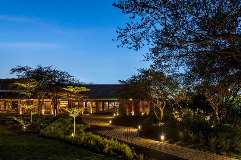 Low-slung pavilion with corrugated roof and timber screen walls along brick paved path through landscaped garden at dusk