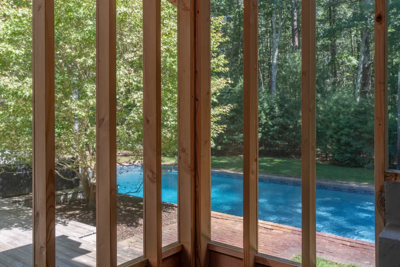 Interior view through vertical timber slats framing a blue pool and wooded garden beyond
