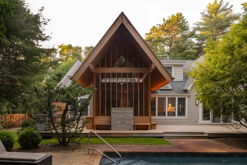 Timber pavilion with steep gabled roof and vertical slat facade beside a swimming pool at dusk