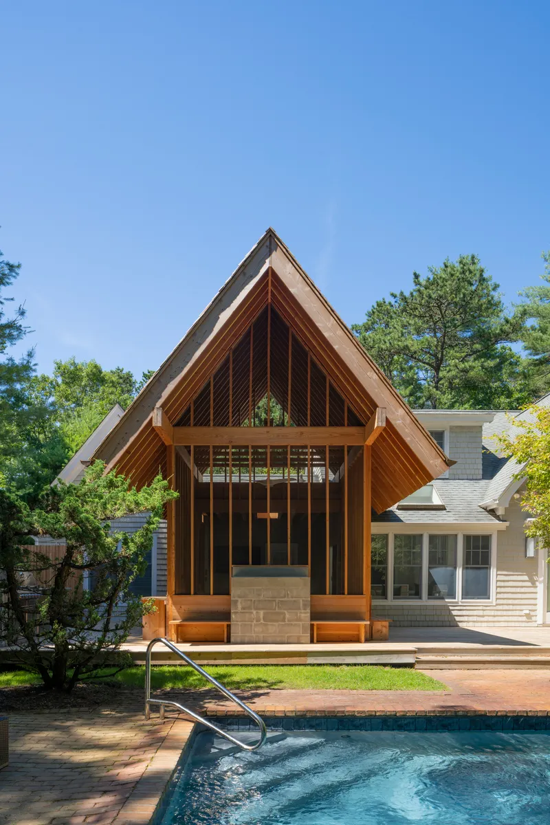 Timber-framed pavilion with steep gable and vertical slats overlooking a swimming pool on a summer day