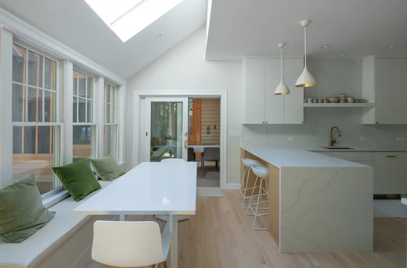 White kitchen with island and pendant lights adjacent to a window seat flooded with daylight