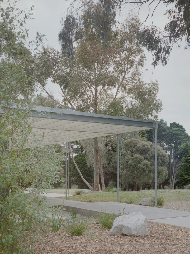 Freestanding pavilion with paneled canopy on steel supports amid grasses and boulders under overcast sky