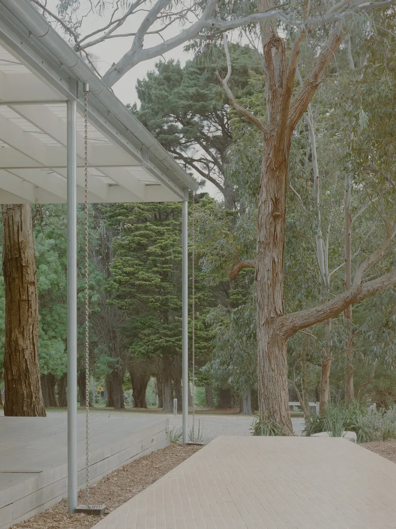 Covered walkway with slender steel columns alongside a paved path surrounded by tall eucalyptus and pine trees