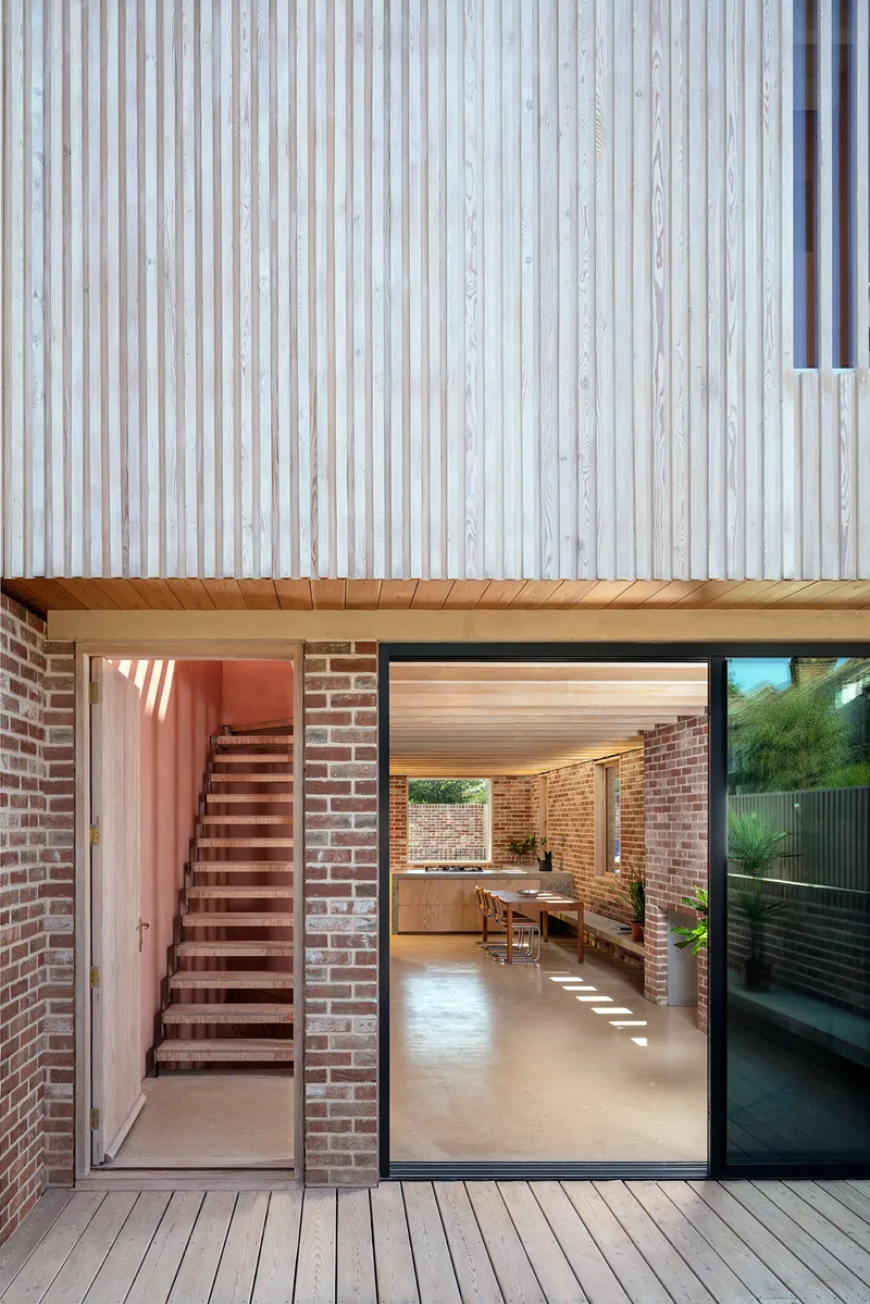 Glazed opening from timber deck to interior with brick staircase visible beneath corrugated metal overhang