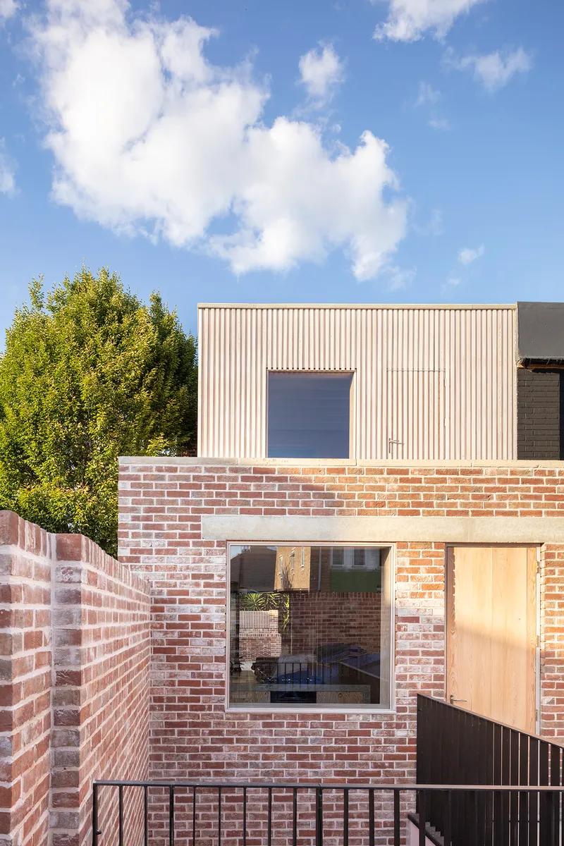 Rear facade with reclaimed brick base and vertical timber cladding above a window under blue sky