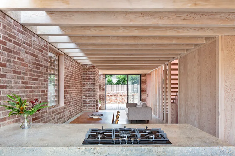 Open kitchen with exposed timber joists above concrete counter and gas cooktop facing brick walls
