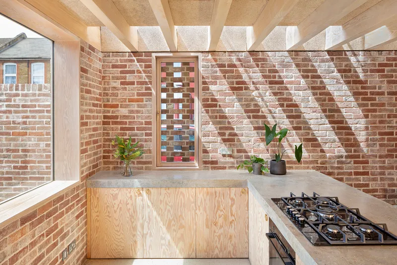 Kitchen corner with plywood cabinetry and slotted vertical window casting diagonal shadows across brick wall