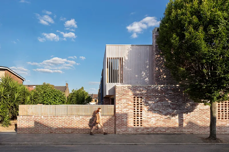 Street view of perforated brick wall with timber fence and corrugated metal upper volume as a person walks past