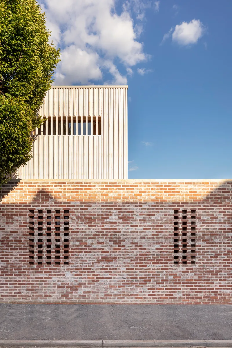 Detail of perforated brick boundary wall with vertical slot openings beneath corrugated metal facade and blue sky