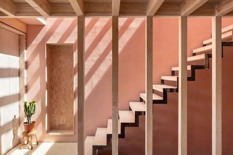 Floating timber staircase against a pink rendered wall with exposed ceiling beams and afternoon sunlight