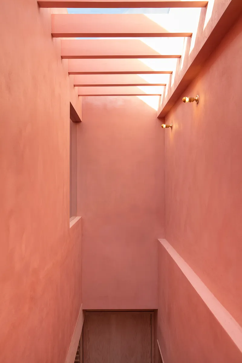 Narrow pink plastered stairwell with horizontal timber ceiling slats and brass wall sconces