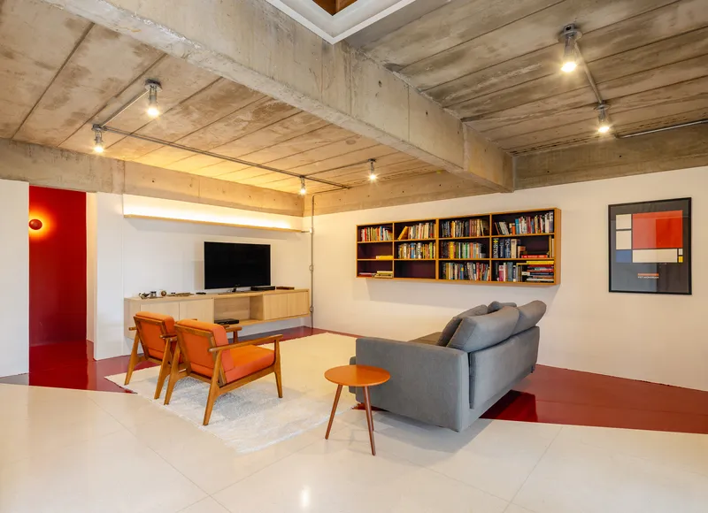 Living room with exposed concrete ceiling, wall-mounted bookshelves, and red resin floor panels under track lighting