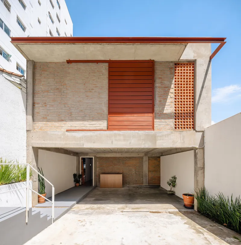 Two-story facade with exposed concrete, brick walls, sliding orange timber panels and carport below under clear sky