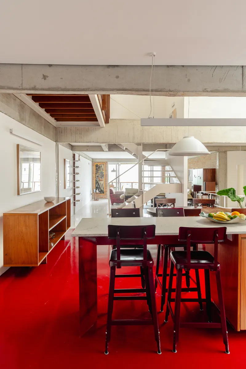 Open plan interior with exposed concrete beams, timber furniture, and bright red floor under pendant lights