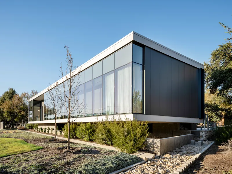 Cantilevered dark metal panel volume over planted beds with young trees and evergreen shrubs below