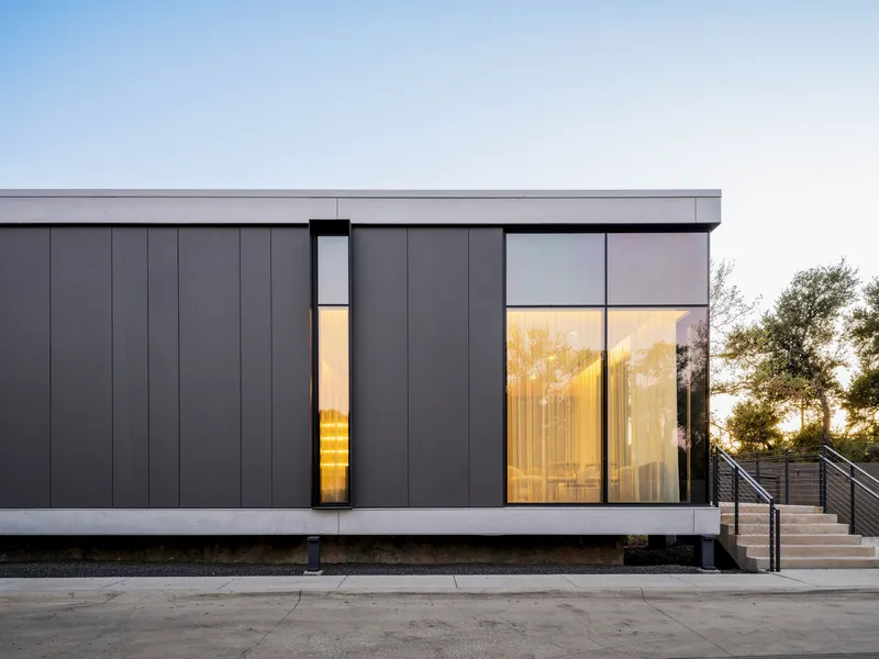 Dark metal panel facade with lit vertical slot window and larger glazed opening at dusk