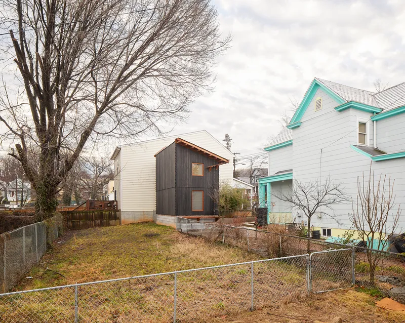 Side view of the compact timber volume on a sloped lot between neighboring clapboard houses