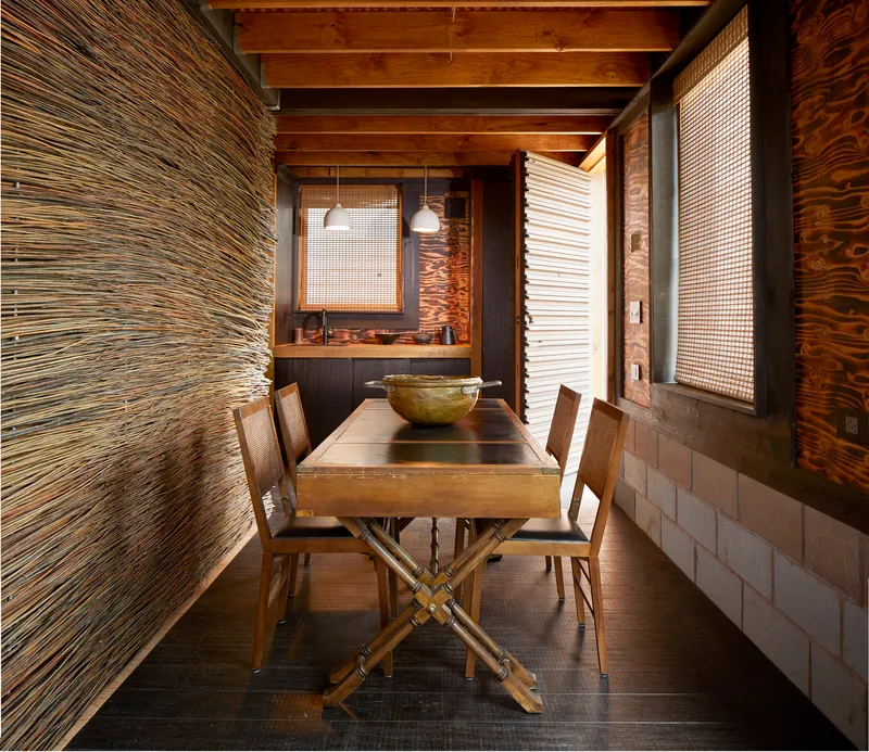 Dining area with exposed timber beams and layered wall treatments of woven screens and plywood panels