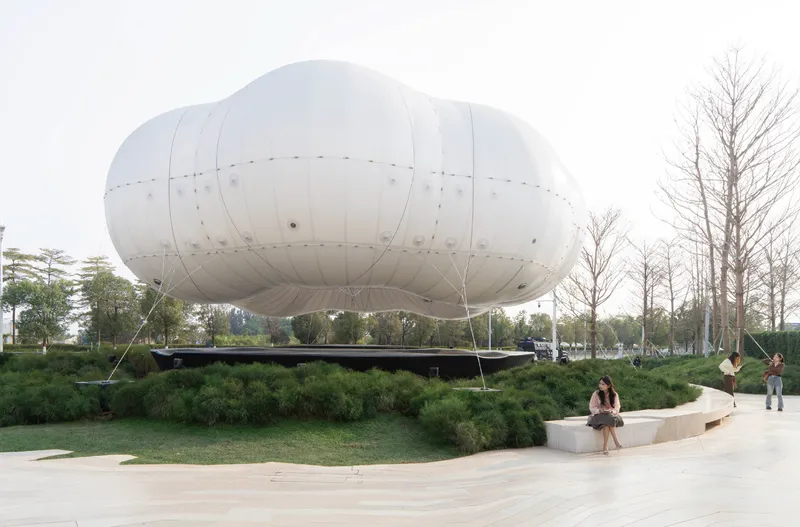 White inflatable balloon structure tethered above planted berms with visitors seated on concrete benches below