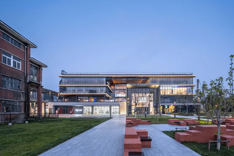 Glazed multi-level building with illuminated interiors facing a courtyard with red brick seating elements at twilight