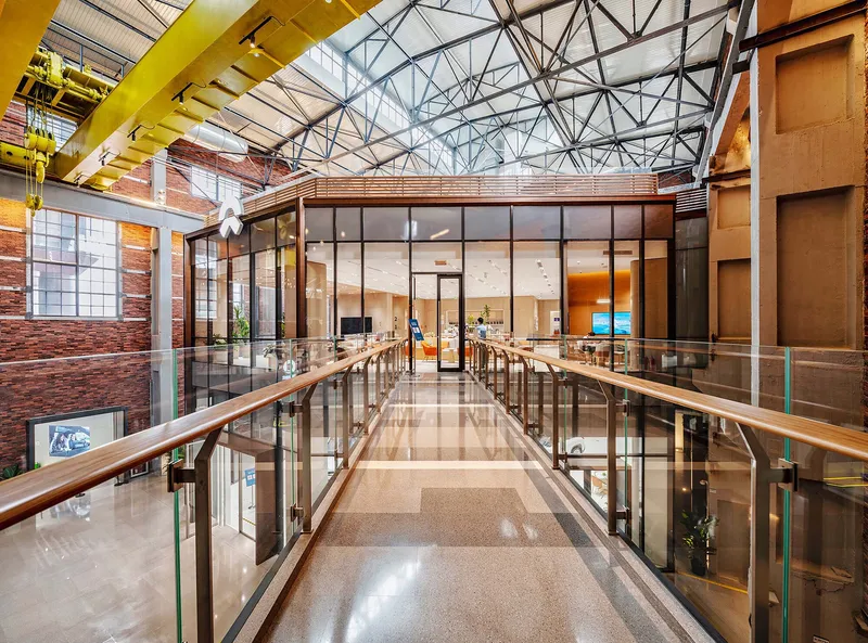 Glass-enclosed pedestrian bridge with exposed steel trusses spanning between brick and timber-clad volumes