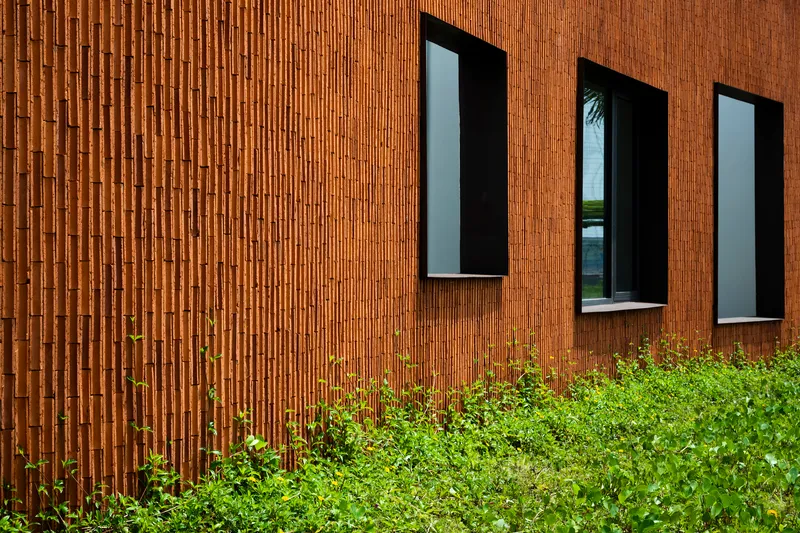 Three tall narrow windows puncturing the vertical terracotta brick facade above green shrubs