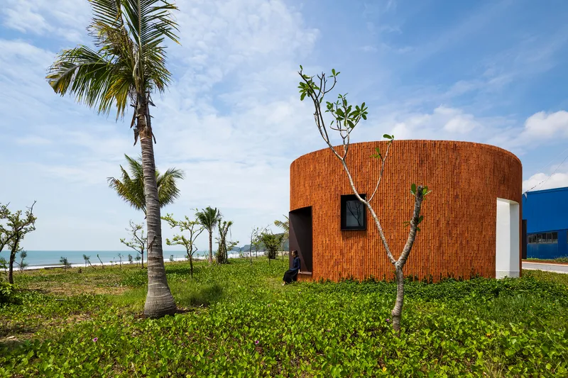 Curved brick volume with small window and door openings amid palm trees and coastal vegetation