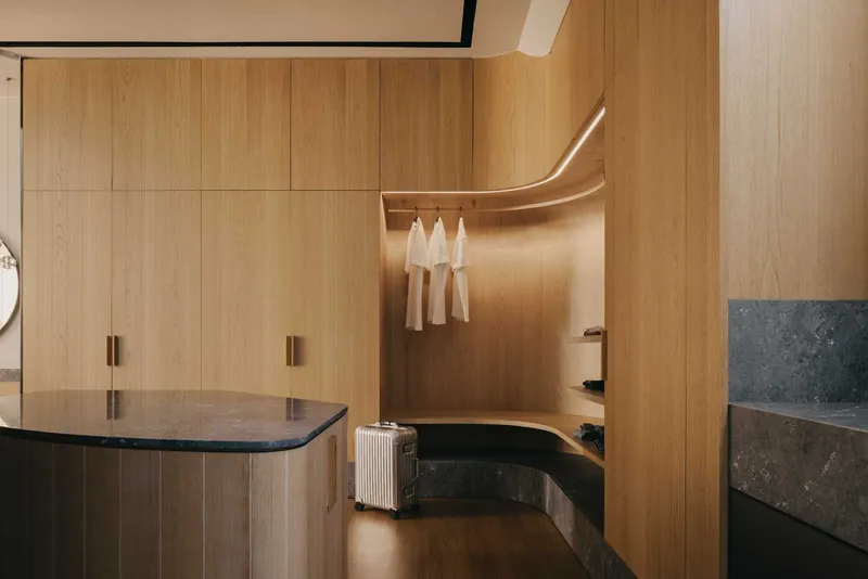 Wardrobe with curved timber hanging rail, integrated lighting, and stone bench seating beside a suitcase