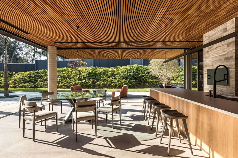 Outdoor dining area beneath timber slatted ceiling with views to manicured hedge and lawn through floor-to-ceiling glass