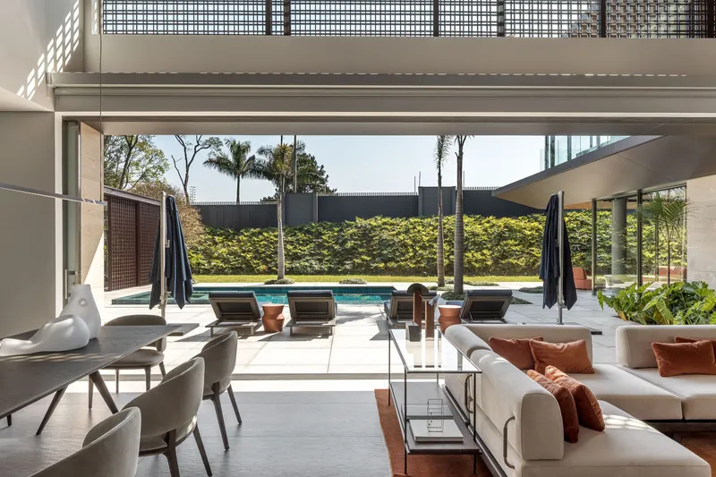 Double-height living room opening to pool terrace with palm trees and hedge wall beyond